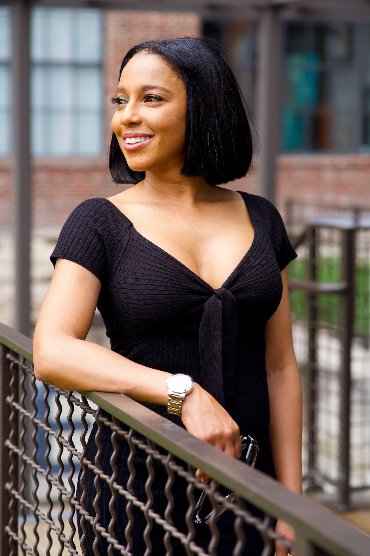 Person in a black dress leaning on a railing with a watch on wrist and holding sunglasses.