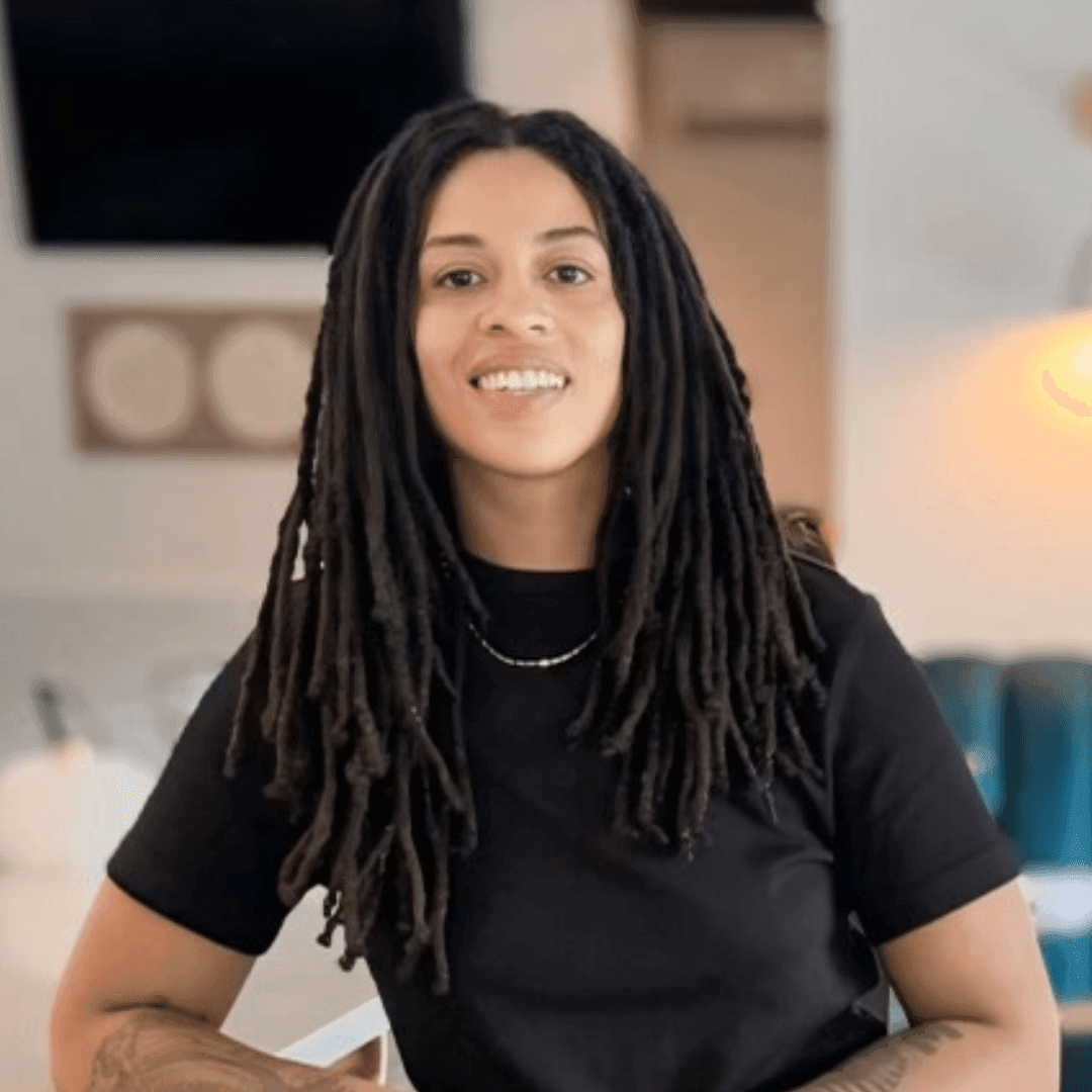 Smiling person with long dreadlocks wearing a black shirt in a modern interior setting.